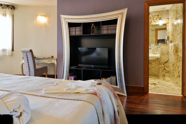 Une chambre d'hôtel élégante avec un lit, un bureau, une télévision et une salle de bains en marbre luxueuse visible à travers une porte ouverte.