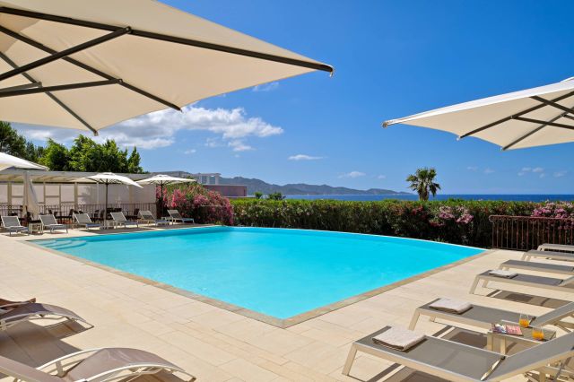 Une scène sereine au bord de la piscine avec des parasols, des chaises longues, des fleurs éclatantes, et une vue sur l'océan et les montagnes.