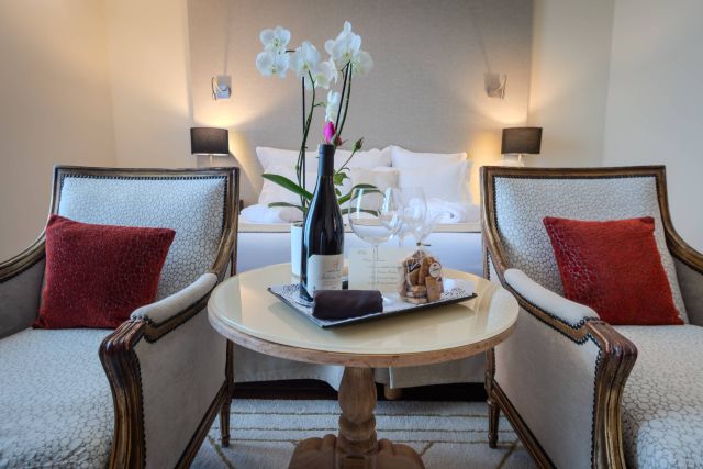 Chambre élégante avec deux chaises, une table garnie de vin, de verres et de snacks, ainsi qu'un lit en arrière-plan et des fleurs.