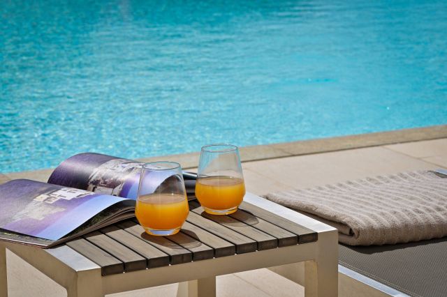 Deux verres de jus d'orange reposent sur une table à côté d'une piscine bleue scintillante, accompagnés d'un magazine et d'une serviette.