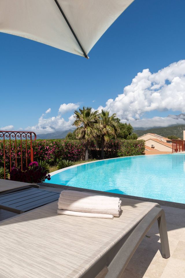 Une scène paisible au bord de la piscine avec un transat, des serviettes, des fleurs éclatantes et un ciel bleu clair. Ambiance de détente.