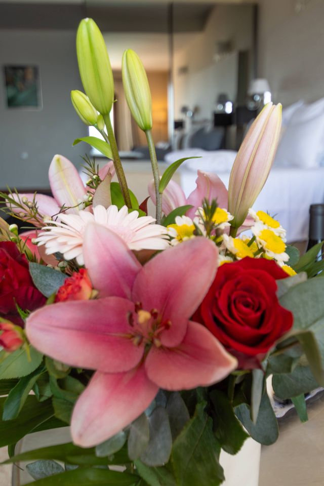 Ein lebhaftes Bouquet mit rosa Lilien, roten Rosen und Gänseblümchen, vor einem unscharfen Hintergrund des Raumes.