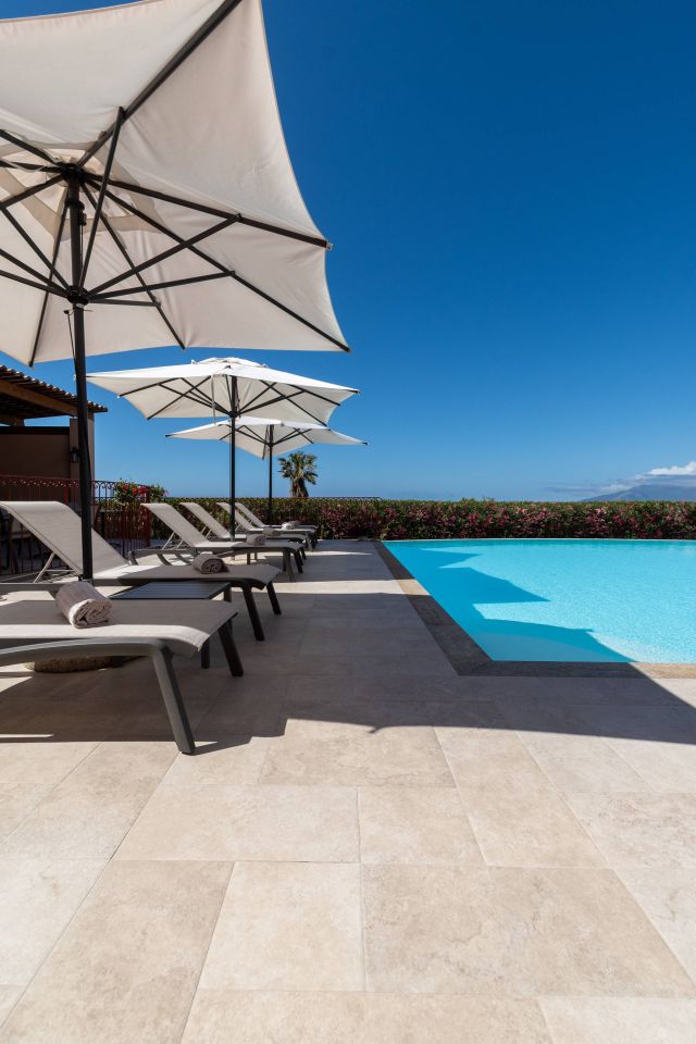 Une scène paisible au bord de la piscine avec des transats, des parasols et un ciel bleu clair, entourée de fleurs vibrantes.