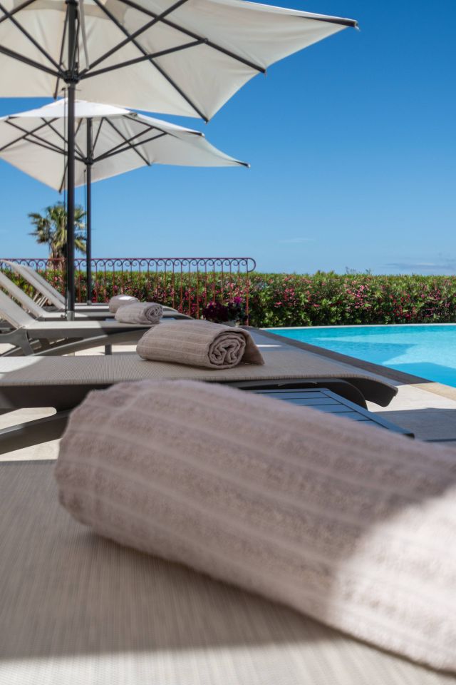 Une scène sereine au bord de la piscine avec des chaises longues, des serviettes enroulées, des parasols et un ciel bleu clair en arrière-plan.