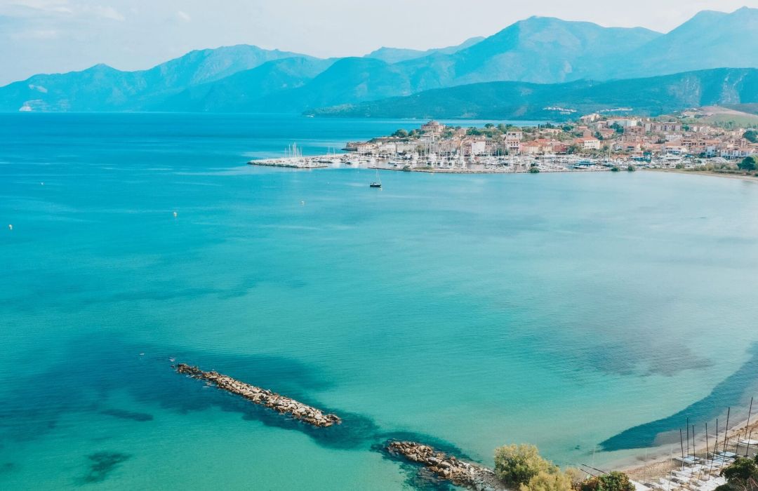Une vue côtière sereine de Saint-Florent avec une eau turquoise claire, des éboulis rocheux et des montagnes lointaines sous un ciel lumineux.