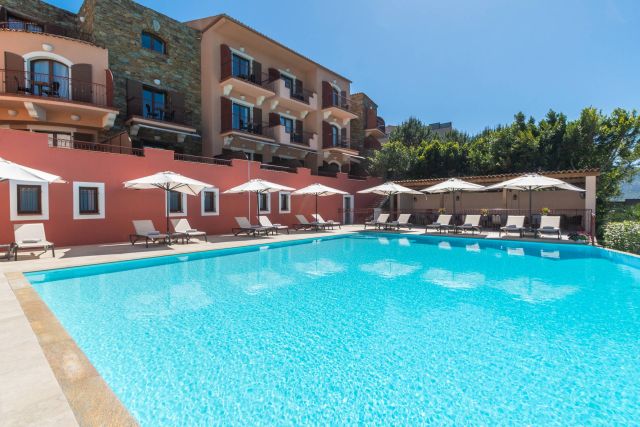 A bright pool area with lounge chairs and umbrellas, set against a colourful hôtel façade under a clear blue sky.
