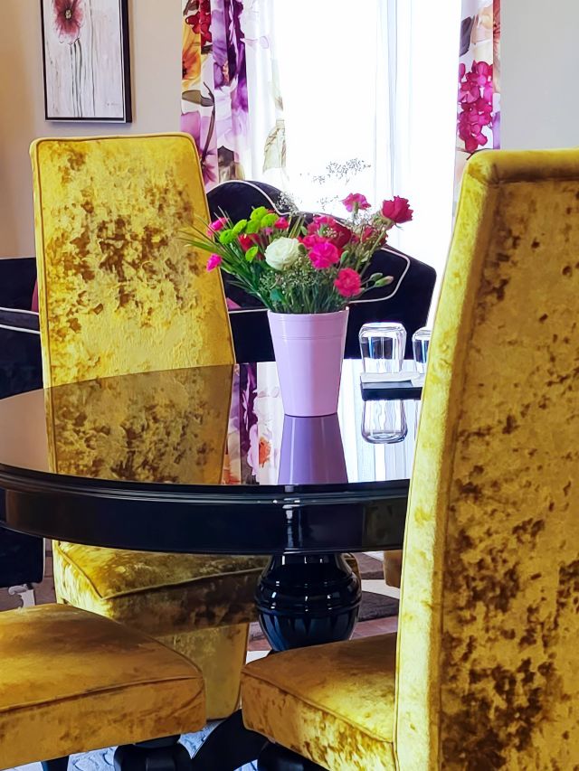 Ein modernes Esszimmer-Setup mit gelben Samtstühlen, einem schwarzen Tisch und einer bunten Blumenanordnung in einer rosa Vase.