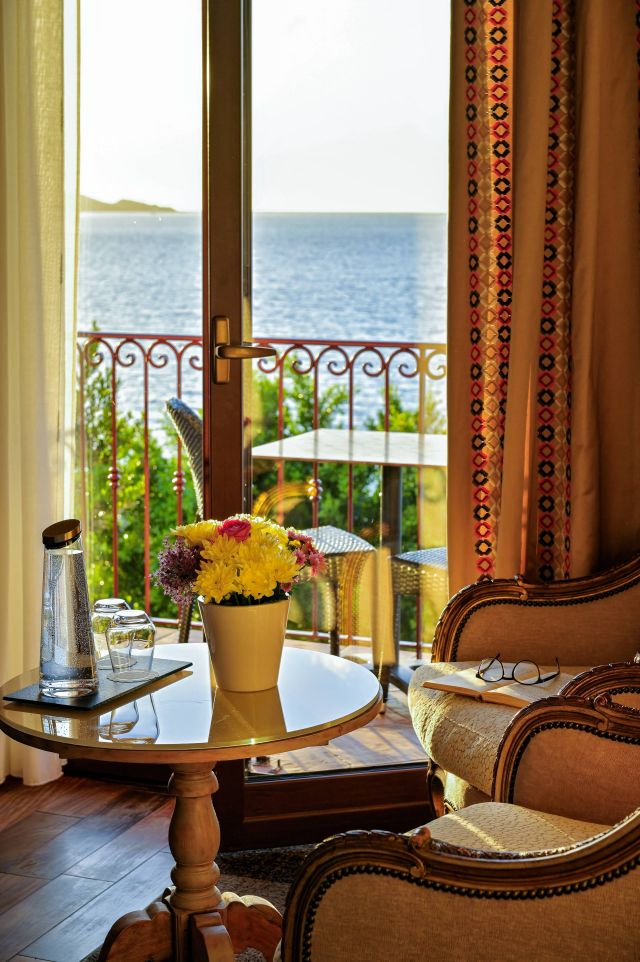 Une chambre cosy avec vue sur la mer, dotée d'une table avec des fleurs, de l'eau et d'une paire de lunettes sur une chaise.