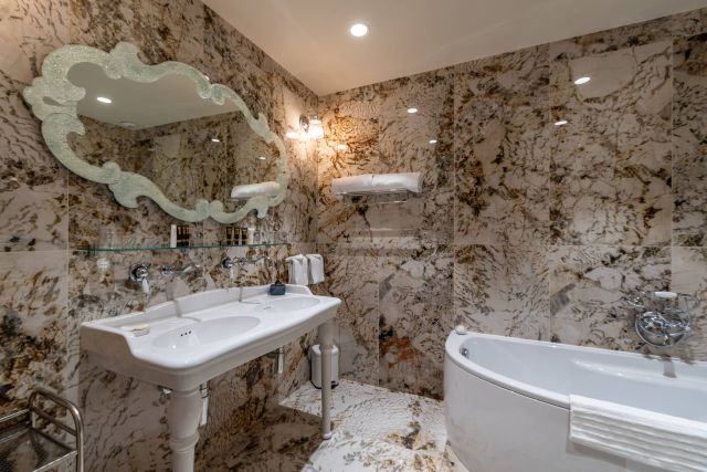 Luxurious bathroom with marble walls, elegant mirror, double sinks, and a bathtub, featuring a sleek design and lighting.