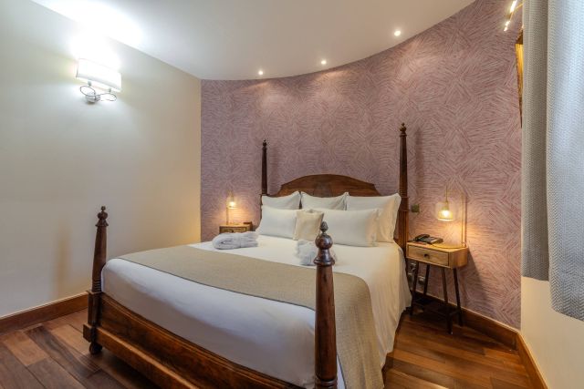 Chambre confortable avec un lit en bois, des draps doux et un éclairage chaleureux contre un mur rose texturé.