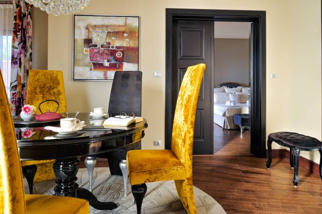 A stylish dining area features a round table, velvet chairs, and a doorway leading to a cosy bedroom.