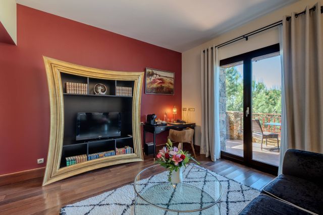 Salon confortable avec une étagère en or unique, une télévision, un bureau et une vue à travers de grandes fenêtres sur un balcon et la verdure.